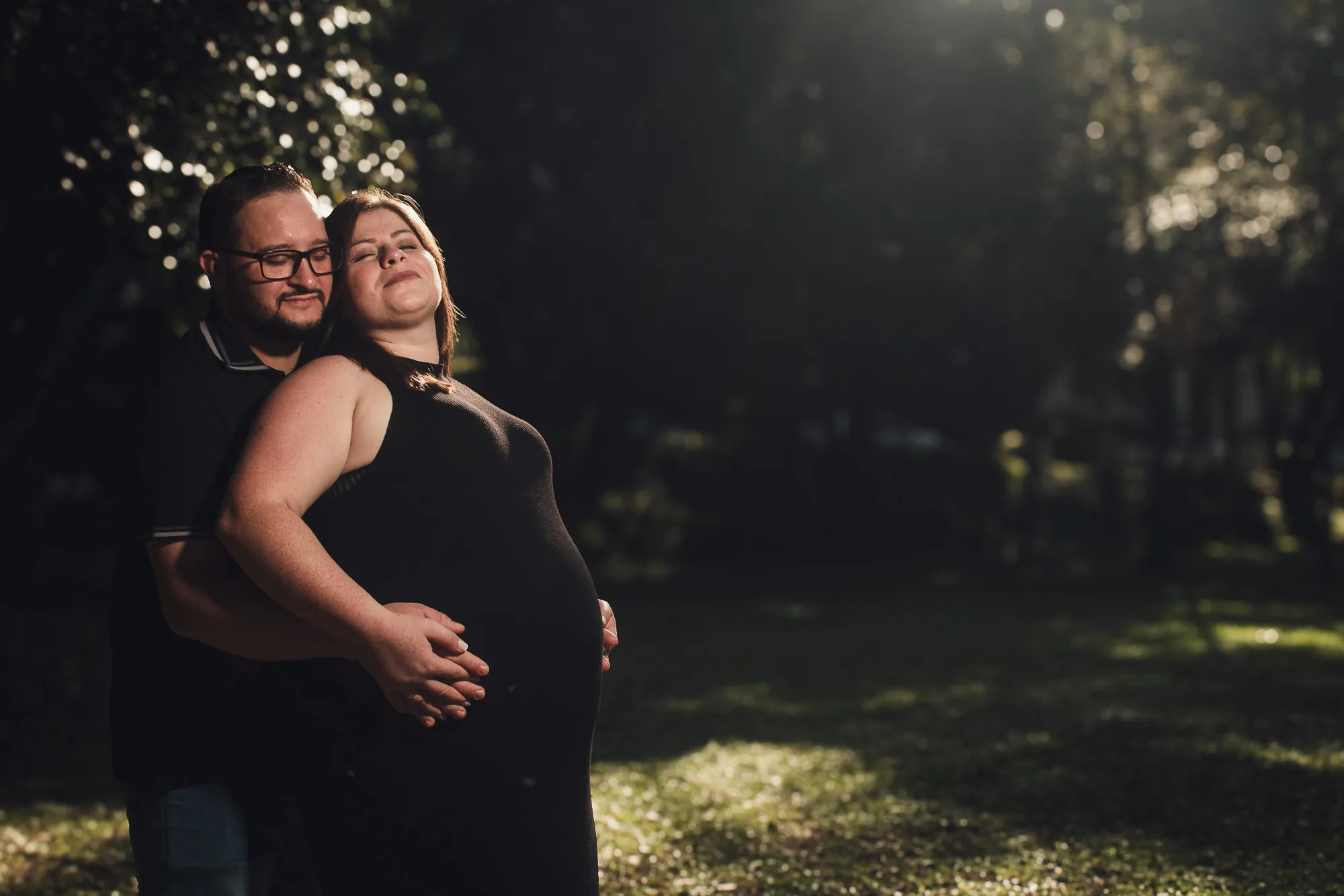 Casal gestante sob a sombra das árvores, mãos na barriga
