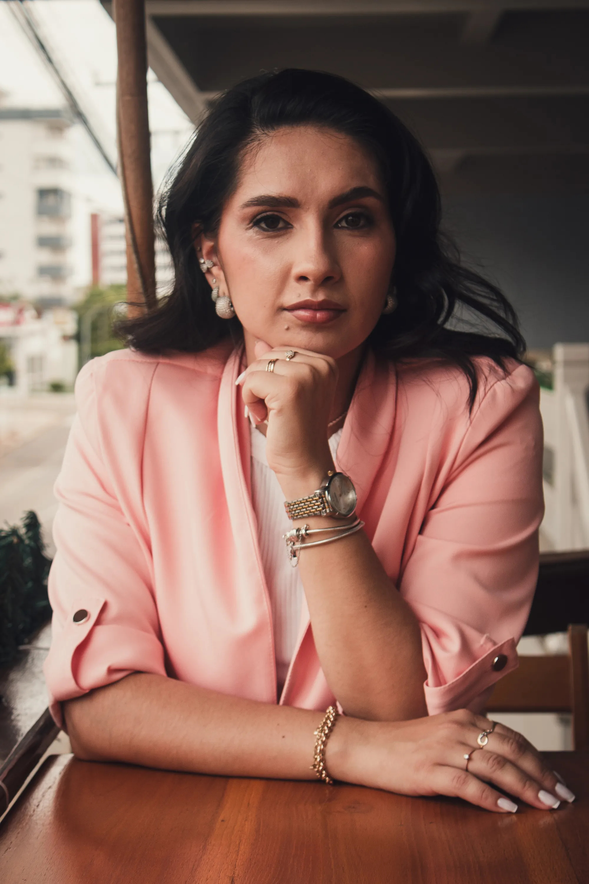 Mulher de blazer rosa, queixo apoiado na mão, em mesa de madeira