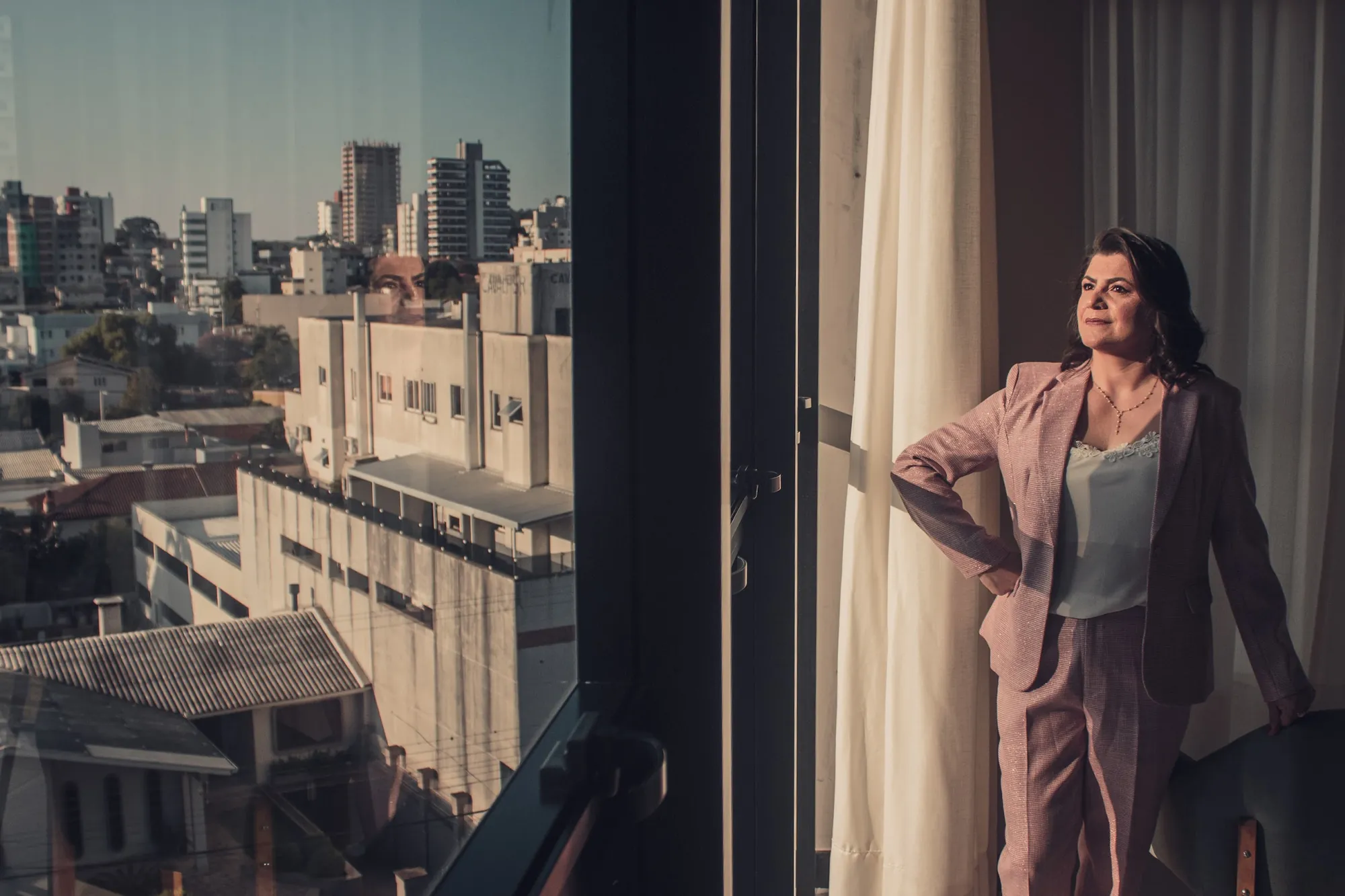 Mulher de terno rosa em janela alta com a cidade ao fundo