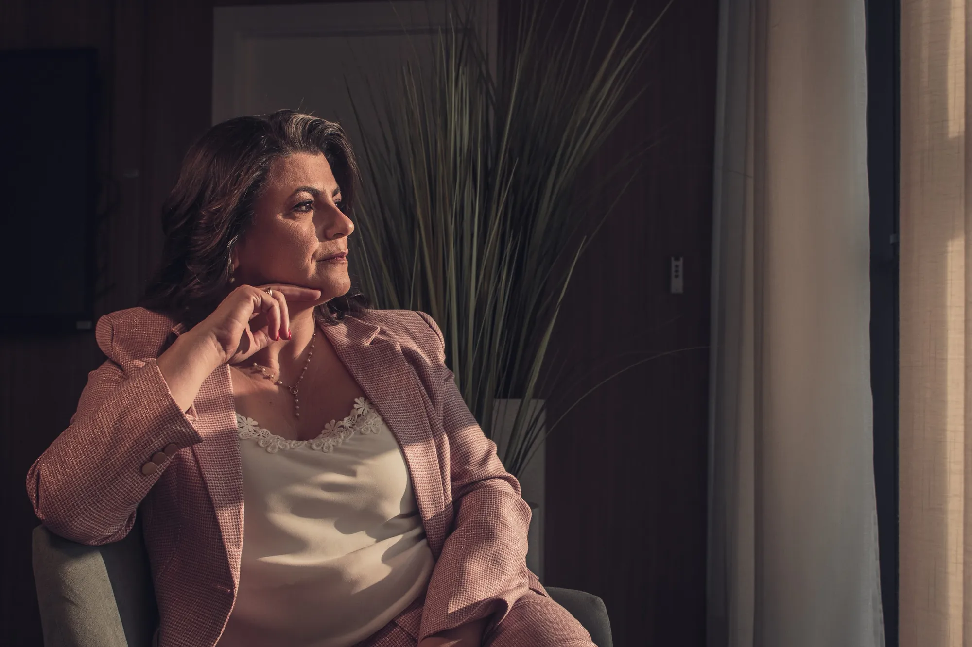 Mulher de terno rosa sentada em poltrona junto à janela