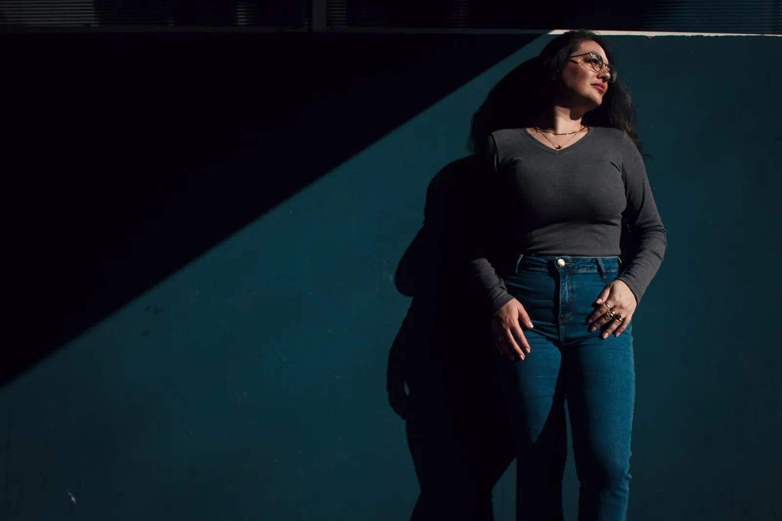 Mulher encostada em parede azul-petróleo cortada por uma diagonal de luz
